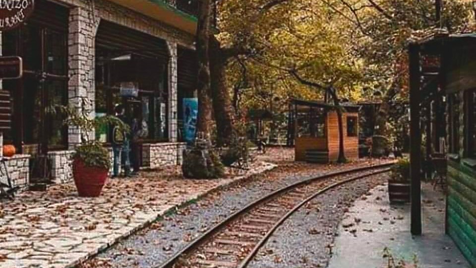 Το Ρομάντζο το φθινόπωρο: η πέτρινη αψίδα και τα τραπέζια κάτω από το κίτρινο πλατάνι, με τις ράγες του Οδοντωτού στρωμένες με φύλλα.